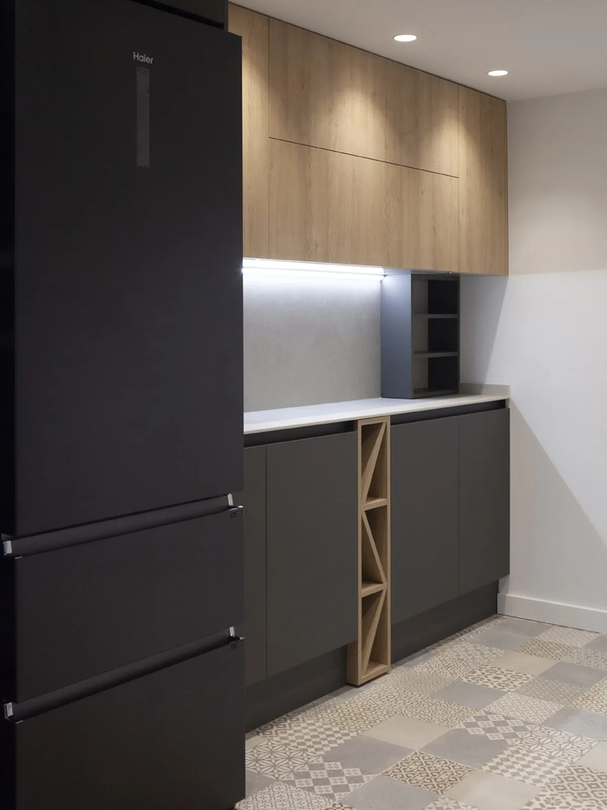La cocina vista desde dentro: el frigorífico, los muebles altos en tono madera y los bajos en gris oscuro, con un botellero de madera entre los módulos