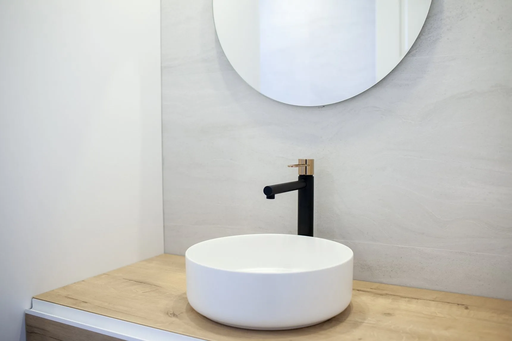 El lavabo redondo sobre la encimera de roble claro, con el espejo redondo y la grifería alta en negro y latón
