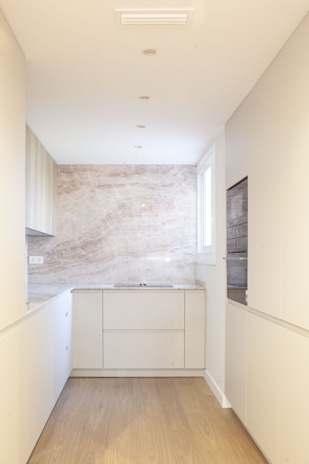 Vista frontal de cocina en U con pared de piedra natural de vetas rosadas, muebles altos en madera gris ceniza y frentes blancos
